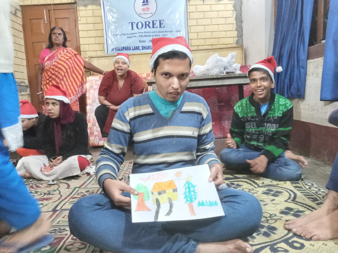 A young man in a Santa hat sitting cross-legged on a mat, holding up his drawing of a house and trees.