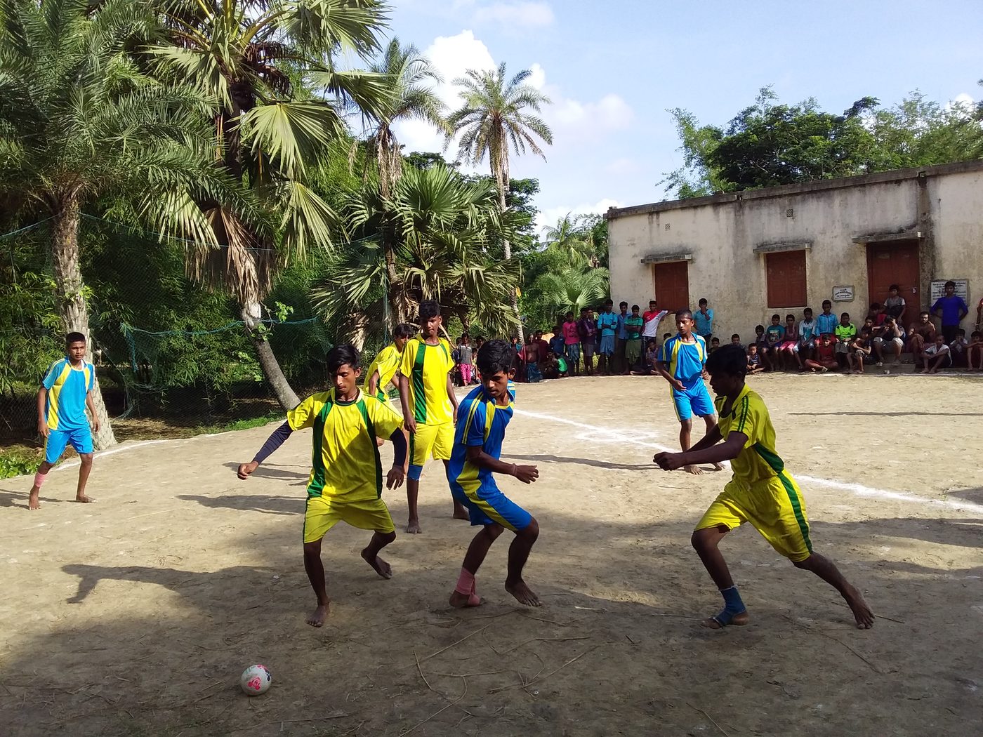 Young players in yellow and blue jerseys contesting the ball on a village ground, watched by a crowd.