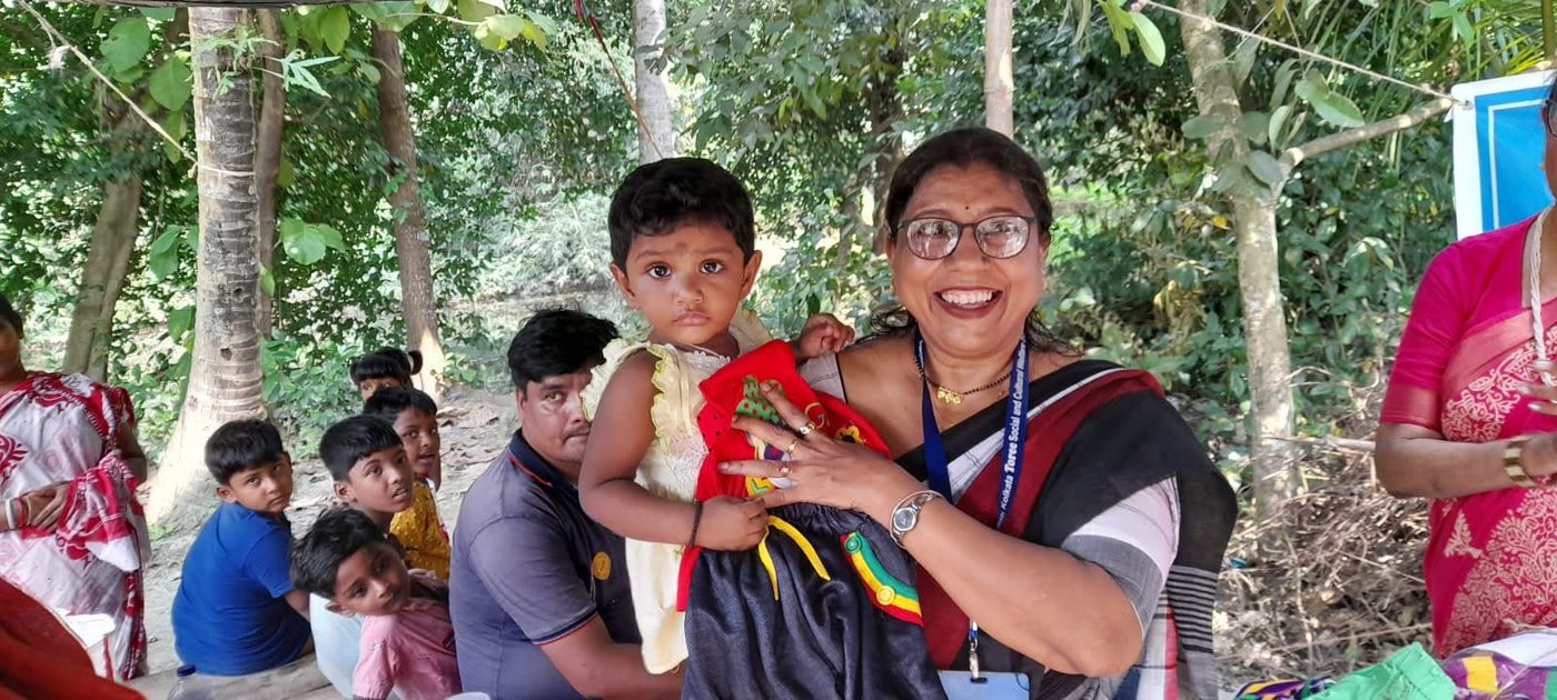 A smiling member of TOREE holding a small child and a set of new clothes at a village camp.