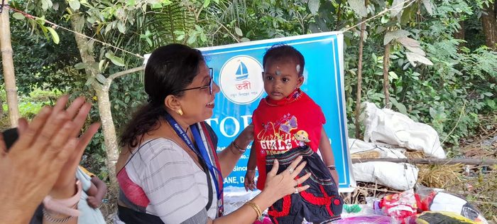 A member of TOREE holding a small child in new clothes beside the TOREE banner, October 2025.