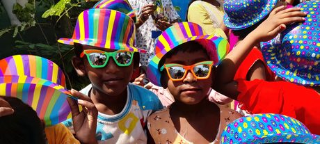 Two village children in party hats and sunglasses at a Holi celebration.