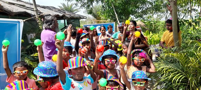 A crowd of children in party hats holding up coloured balls beside a TOREE banner.