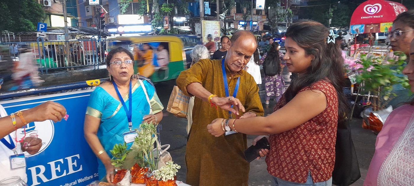 A member of TOREE tying a rakhi on a young woman's wrist at the TOREE stall on a Kolkata street, with potted plants ready to be given as gifts.